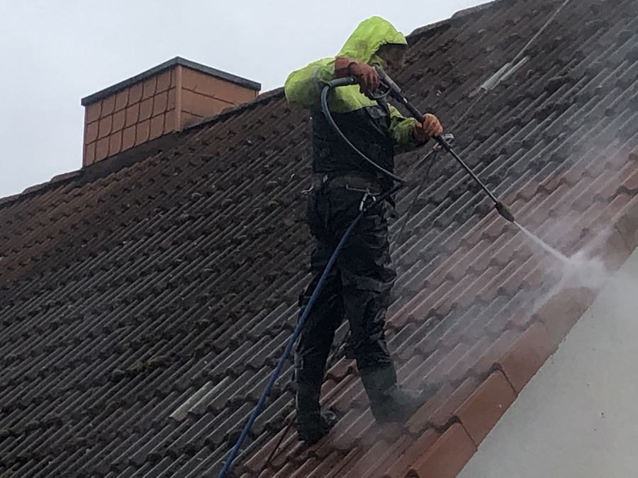 Arbeiter in Schutzkleidung reinigt ein dunkles Ziegeldach mit heißem Wasser, Dampf steigt auf